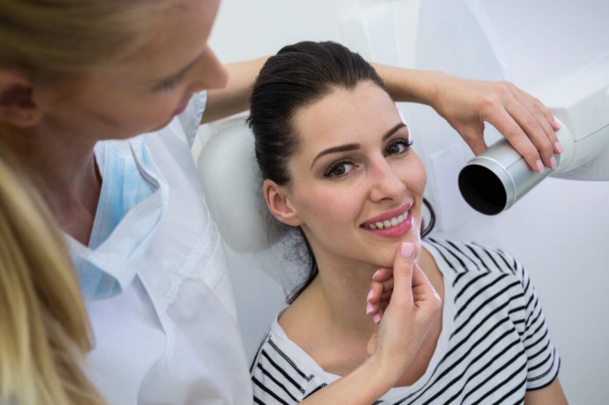 Dentist examining a female patient’s teeth with dental imaging equipment during an in-office oral exam and implant consultation.