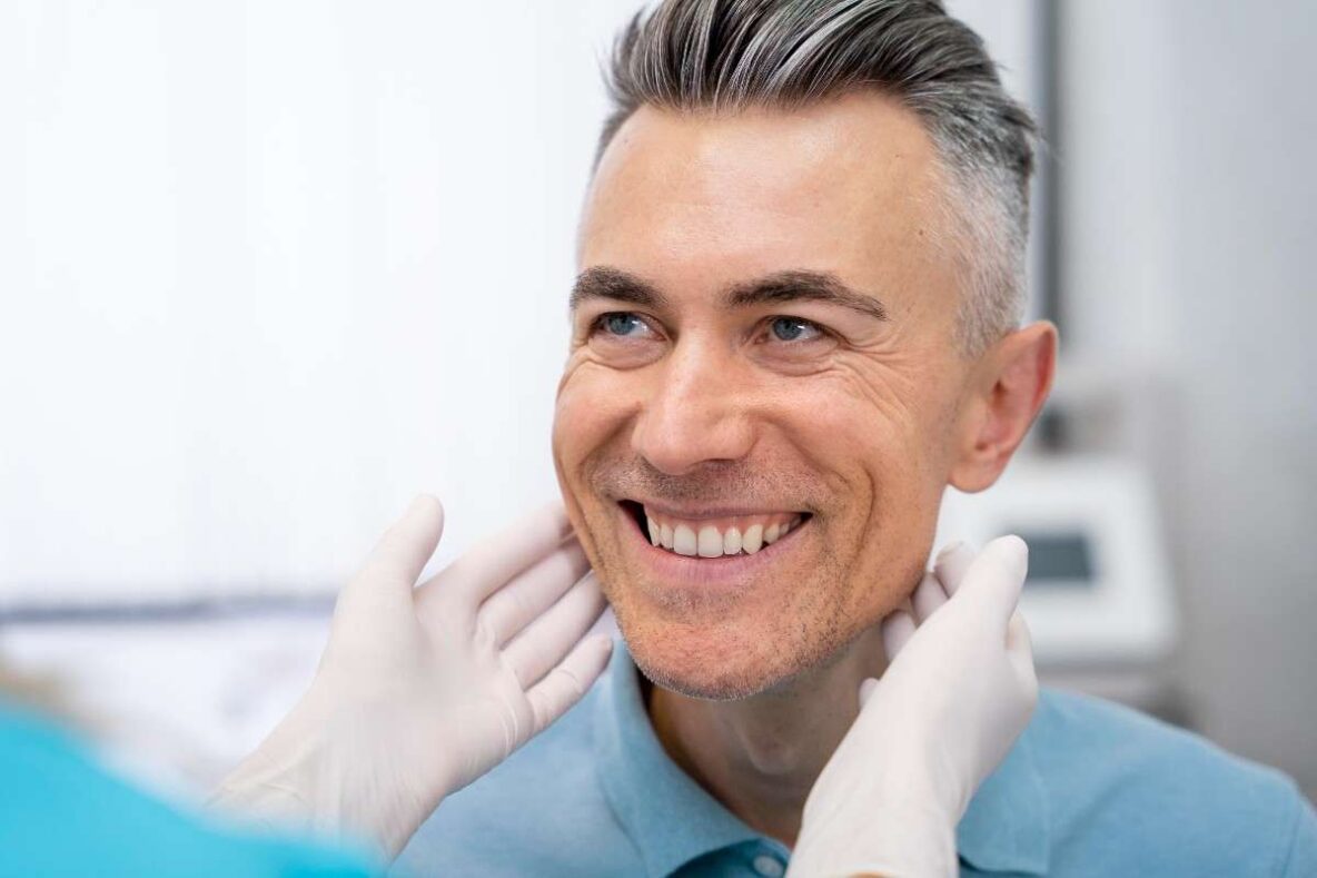 Smiling male patient with a full set of natural-looking dental implants being examined by a dentist wearing gloves in a modern dental clinic.